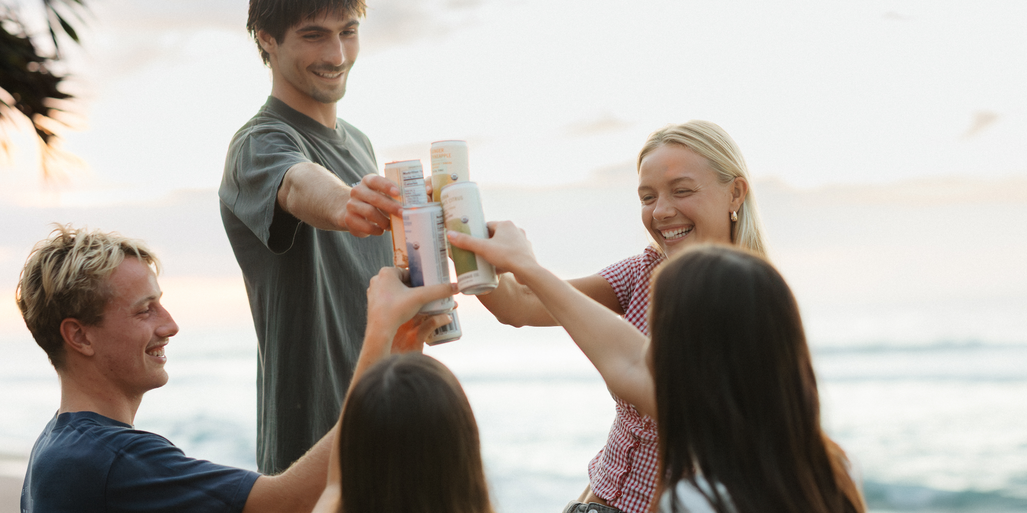 Group of people toasting with cans on a beach at sunset